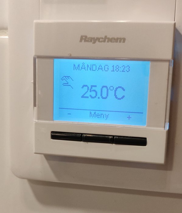 En termostat visar 25°C, måndag 18:23. Två knappar under skärmen, varumärke Raychem.