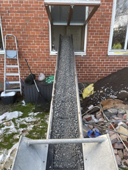 Grusgång under anläggning med skottkärra, stege och trädgårdsredskap vid tegelhus, delvis snötäckt gräsmatta.