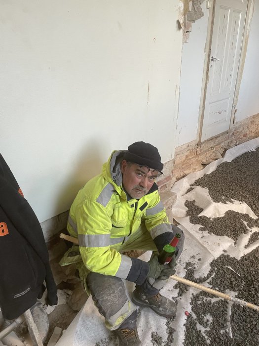 En man i arbetskläder sitter på huk vid en raserad vägg inomhus, med verktyg i handen.