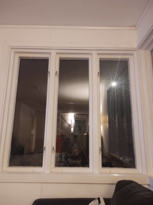 Vit fönsterram med tre glasrutor, speglad inomhusmiljö, oren på högra rutan, del av soffa synlig.