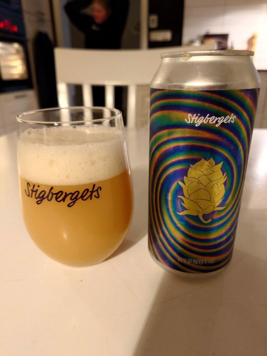 Ölburk från Stigbergets med regnbågsdesign, ölglas med skum, köksmiljö, suddig person i bakgrunden.