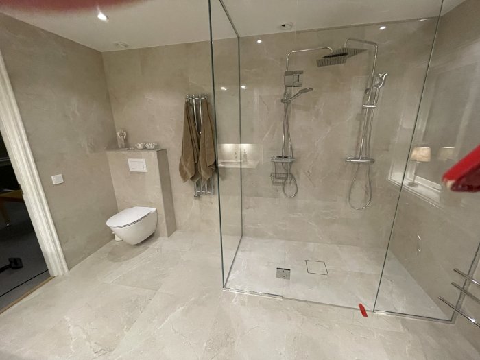 Modernt badrum med dusch, toalett, beige kakel, glasvägg och rostfria detaljer.