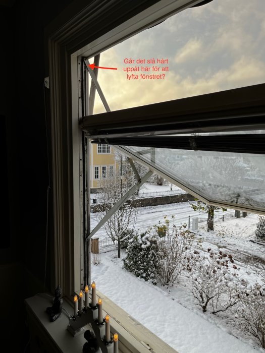 Öppet fönster med utsikt över vinterträdgård, julstjärna, ljusstake och snö.