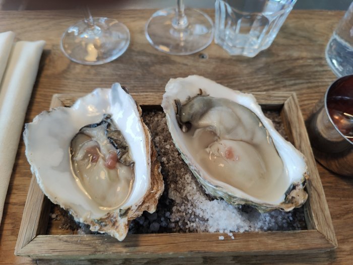 Två ostron på grovsalt, serverade på träbricka, restaurangmiljö med glas och servetter.