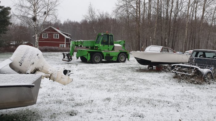 Snöig gräsmatta, grönt fordon, båt på trailer, hus i bakgrunden, träd, grå himmel.