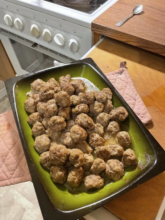 Köttbullar i ugnspanna, vitt kök, spis i bakgrunden, träslev, kökshandduk.