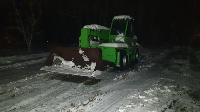 En övergiven grön gaffeltruck med skopa, snötäckt nattetid i en skogsmiljö.