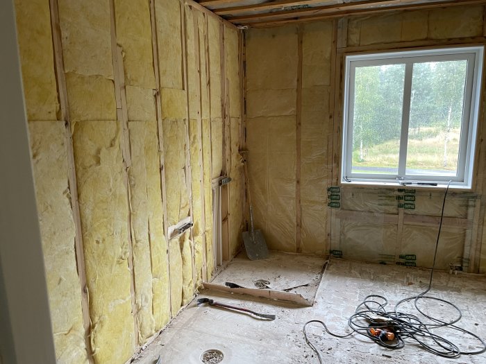 Under renovering, isolering exponerad, oavslutade väggar, arbetsyta, sladdar på golvet, fönster med utsikt utomhus.