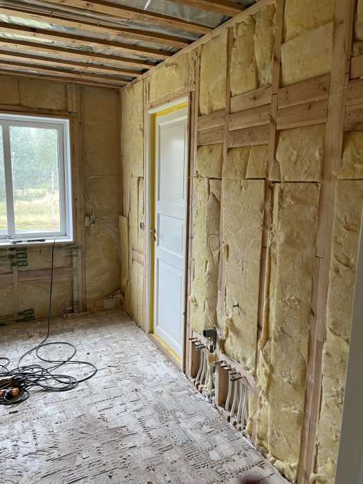 Rum under renovering, isolering synlig, trästomme, obeklädd vägg, fönster, dörr, elinstallation, byggplatskabel på golvet.