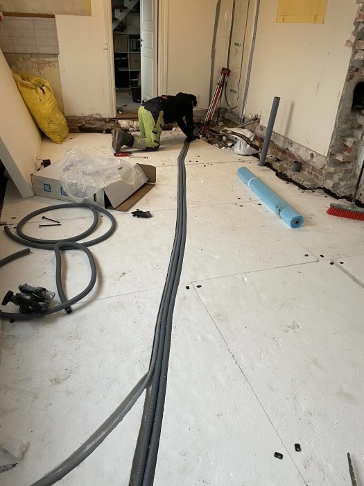 Omfattande renovering inomhus, arbetare böjer sig, golv öppet, rör, kablar, byggmaterial spridda, tegelsten, verktyg synliga.