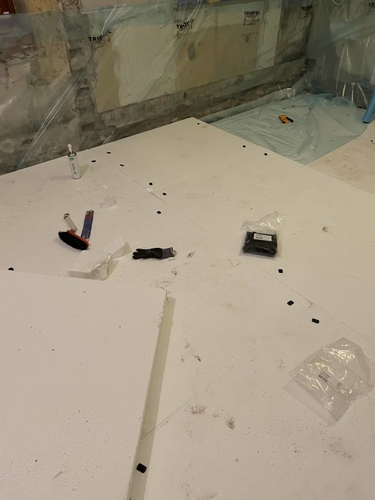Renoveringsplats, isoleringsskumplattor, verktyg, byggspill, skyddsplast, rörbit, osammanhängande inredning.
