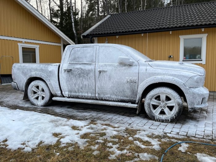 En pickup täckt med skum vid en husfasad under en tvättprocess, med snö på marken.