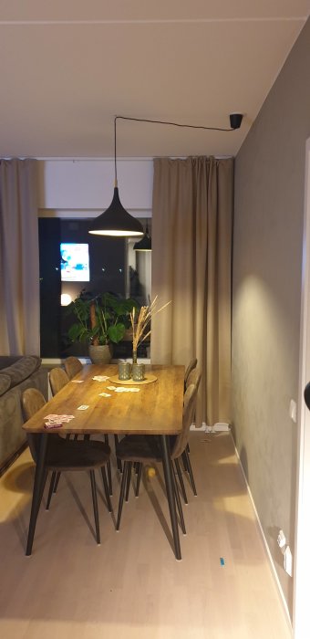 Ett modernt matsalsrum med träbord, stolar, en pendellampa, gardiner och en TV i bakgrunden.