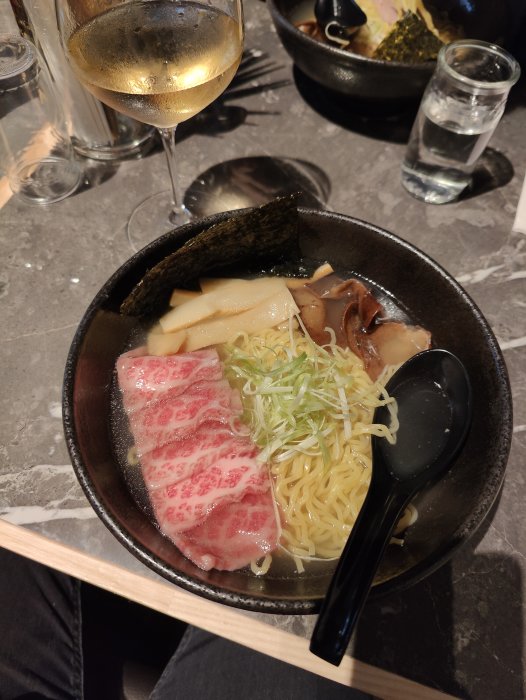 En skål ramen med kött, nudlar och grönsaker, glas vin, vatten, atmosfärisk restaurangmiljö.