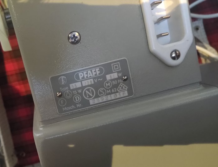 Etikett på en symaskin från PFAFF med specifikationer och modellinformation. Grå bakgrund, metallskruvar och textur.