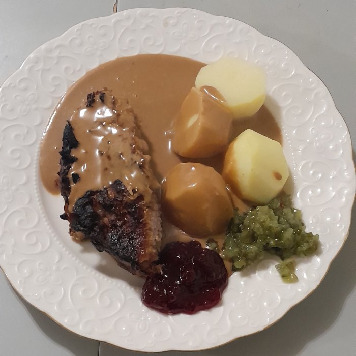 Middagstallrik med kött, sås, potatisar, gelé, rårörda lingon och pressgurka på ett vitt mönstrat fat.