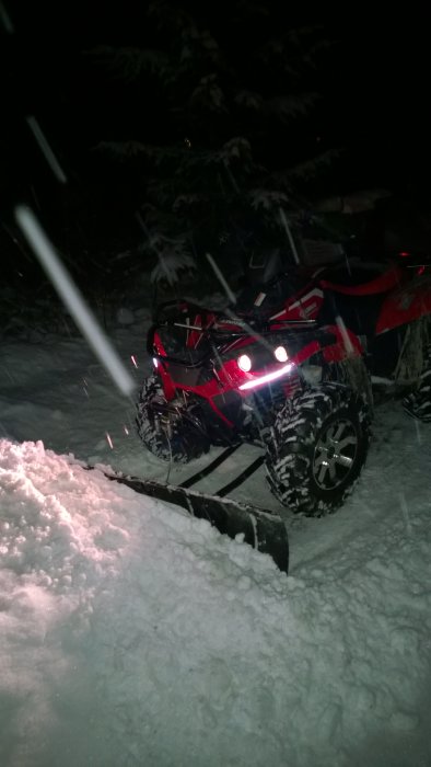 Fyrhjuling med plog i snö, natt, starka lampor, mörk skogsbakgrund, snöfall.