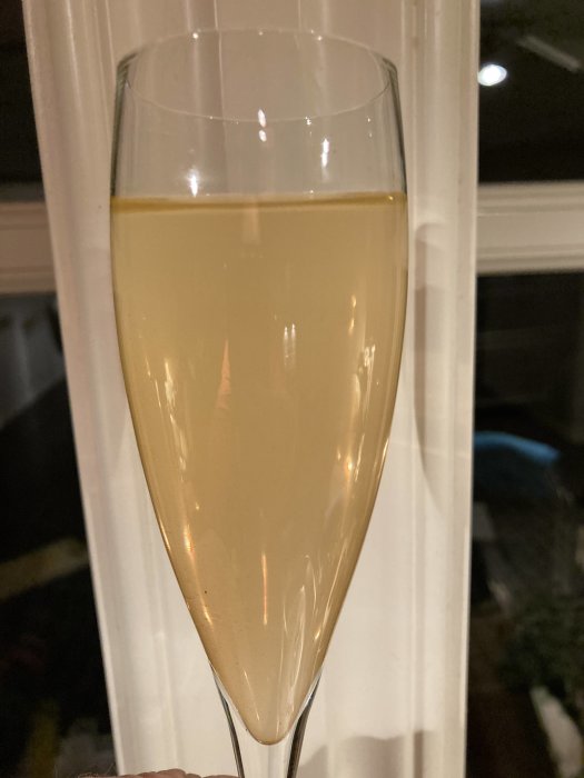 Ett fyllt champagneglas hålls upp, gult skummande innehåll, oskarp bakgrund med fönster.