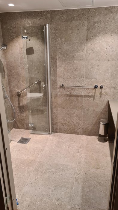 Modernt badrum, duschutrymme med glasdörr, kaklade väggar och golv, handfat, belysning ovanför.