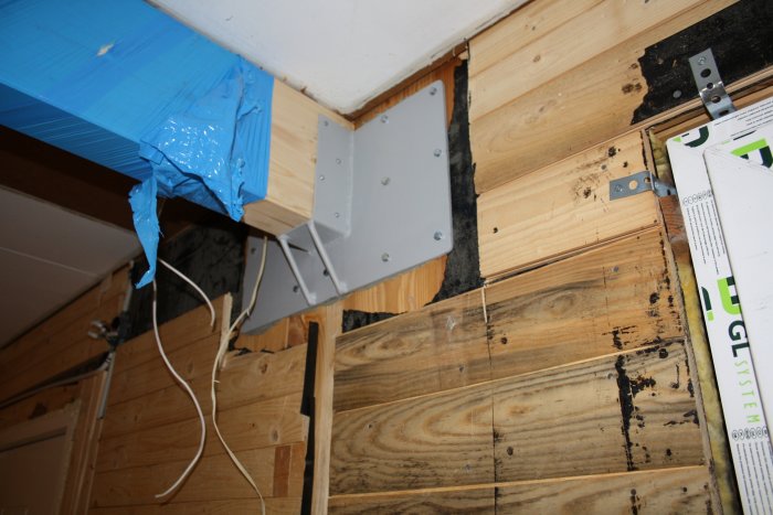 Wooden wall structure with a metal bracket supporting a beam. Blue plastic cover on beam, visible screws, and wiring against bare wall planks.