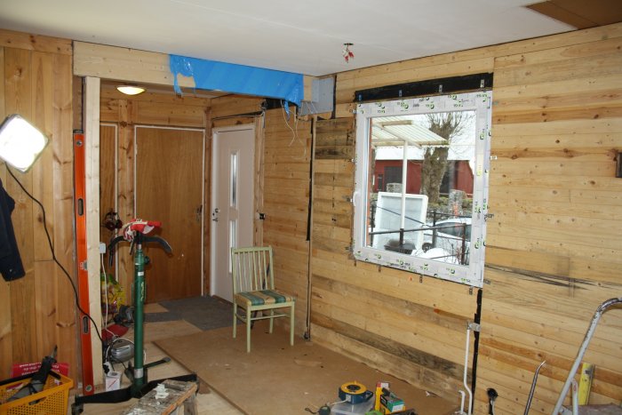 Interior renovation scene with wooden walls, an open window frame, tools scattered, a green chair, and a ceiling beam installation.