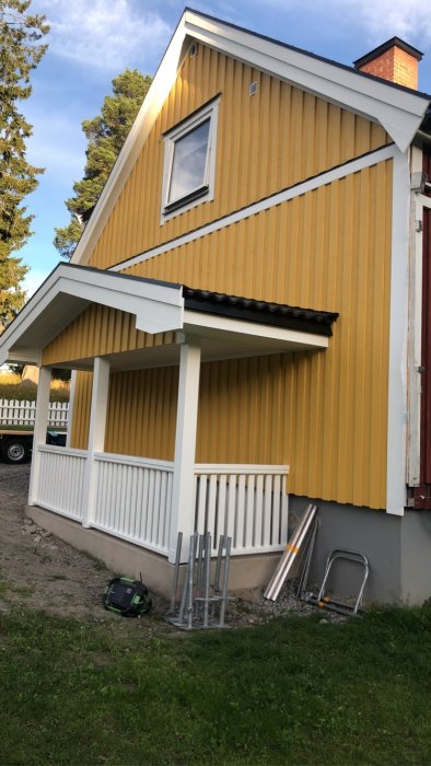 Gult hus med veranda, byggmaterial utanför, grön gräsmatta, klart väder.