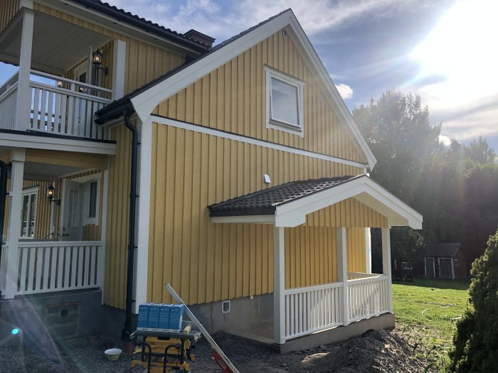 Gult hus med veranda, balkong, solsken, byggmaterial, grönska, friliggande uthus i bakgrunden.