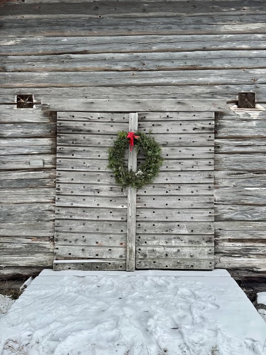 En dörr till lada med julkrans, röd rosett, vit snö framför och åldrat trä.