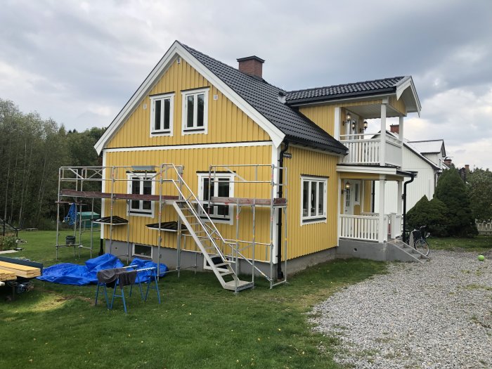 Gult hus med vit veranda, byggställningar, cykel, gräsmatta och grusgång.