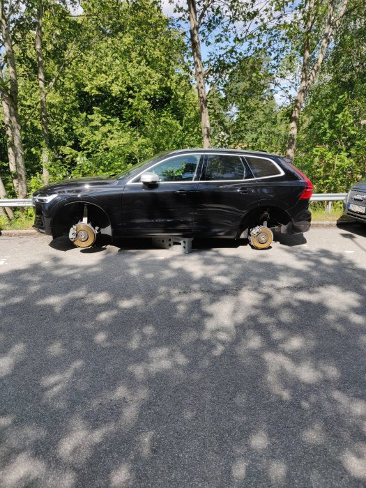 Svart bil utan däck parkerad vid vägkanten, stödd på gula slangar, i dagljus med träd bakom.