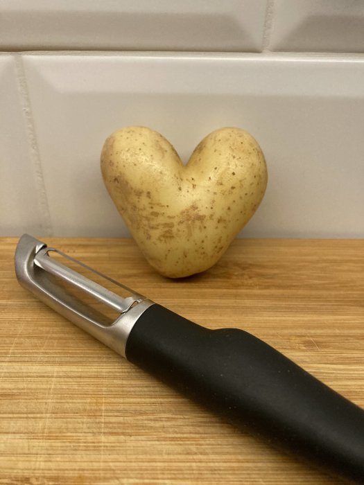 Potatis formad som ett hjärta lutad mot en kaklad vägg, bredvid en svart potatisskalare på ett träskärbräde.