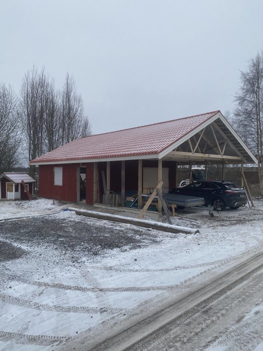 Halvfärdig byggnad, röd med tak, bil inne, snö på mark, stödställningar, grå himmel, träd i bakgrunden.