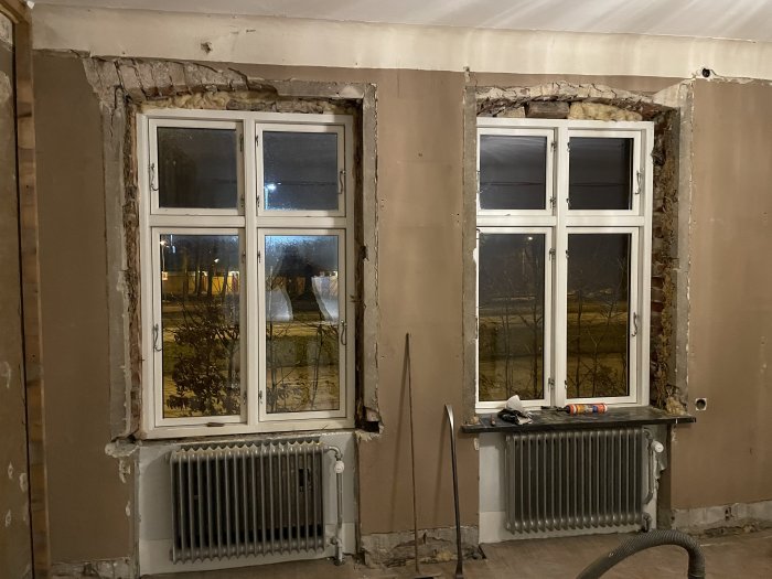 Två fönster med radiatorer nedanför, i ett rum under renovering. Synliga tegelväggar och isolering, nattutsikt utomhus.
