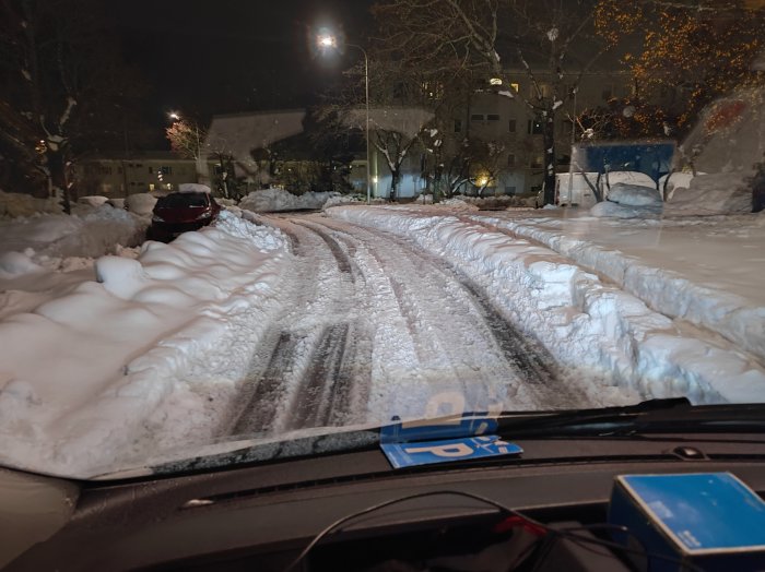 Snötäckt gata på natten, spår av fordon, parkerade bilar, belysning, sett från bilens förarsida.