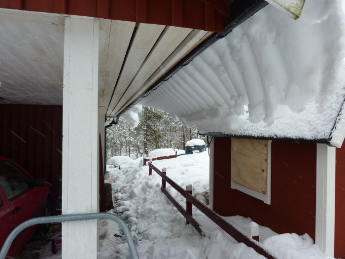 Taket med snö hänger över kanten nära ett bostadshus, vägg med fönster, snötäckt omgivning.