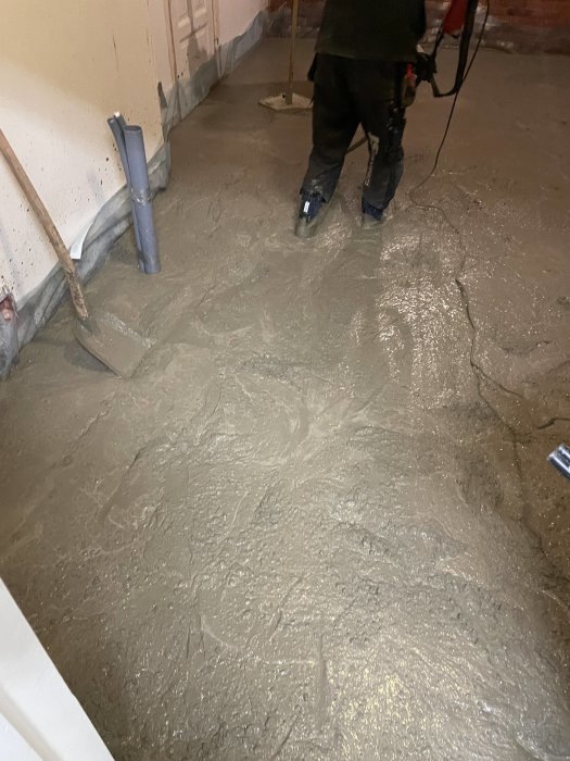 En person jämnar ut nylagt, vått golvbruk i en ofärdig inomhusmiljö. Verktyg och kablar synliga.