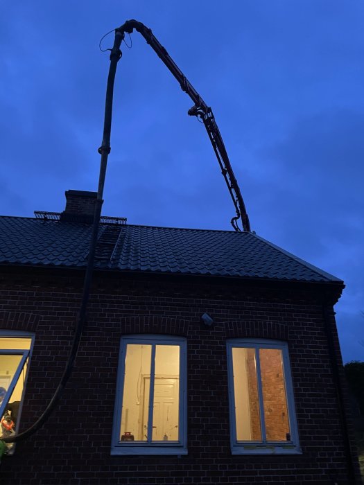 En betongpump med utsträckt arm över ett tegelhus i skymningen.