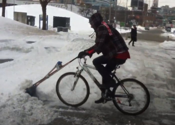 Cyklist i vinterkläder plogar snö med anpassad cykel och skovel. Kallt, urbant landskap.