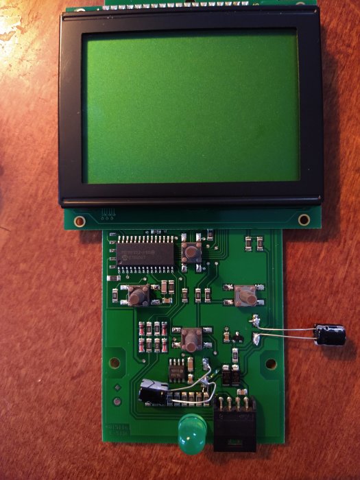 Elektronisk kretskort med komponenter och ett grönt LCD-display ovanpå på träbord.