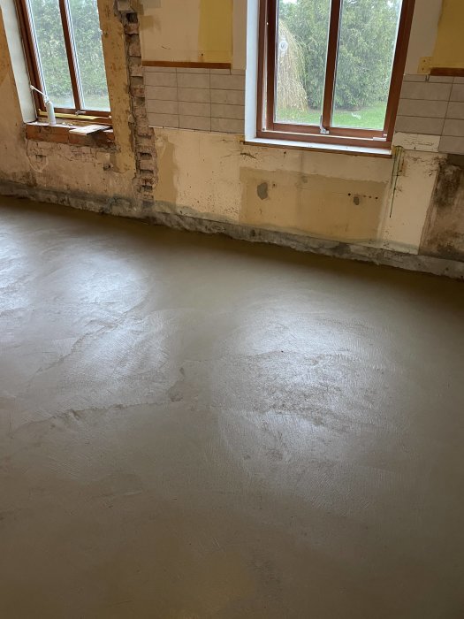 Renoverat rum med nygjuten betonggolv, väggar delvis kala, fönster med utsikt mot grönska.