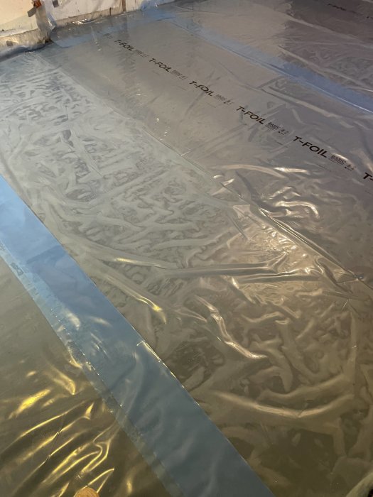 Grå plastfolie på golv, tejpade skarvar, rynkor och veck, textmärkt.