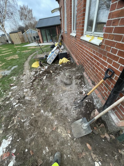 Rörig trädgård, grävarbete, tegelvägg, trädgårdsverktyg, skottkärra, oorganiserat byggavfall, påbörjat projekt.