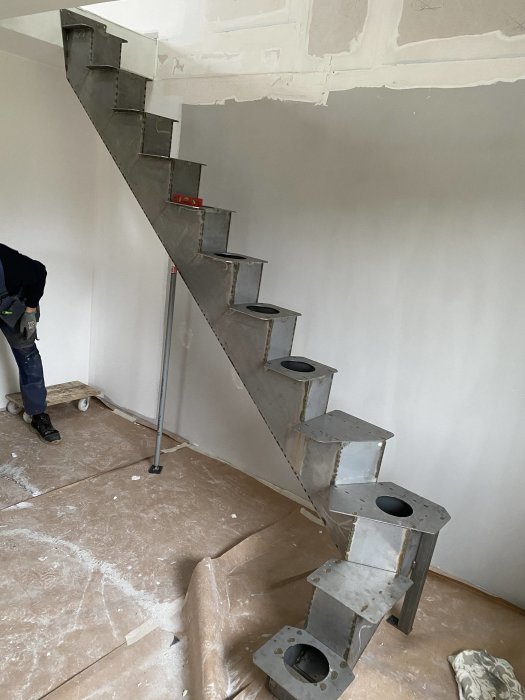 Modern spiral staircase under construction in a room, protective covering on floor, person's arm visible.