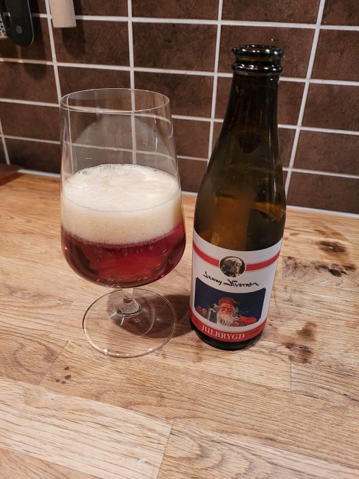 Ett glas med öl och en tom ölflaska på träbänk, kakelvägg i bakgrunden.