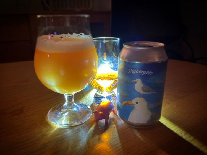 Ölglas med skum, burk med fågeldesign, whiskyglas, varmt ljus, skuggor, träbord.