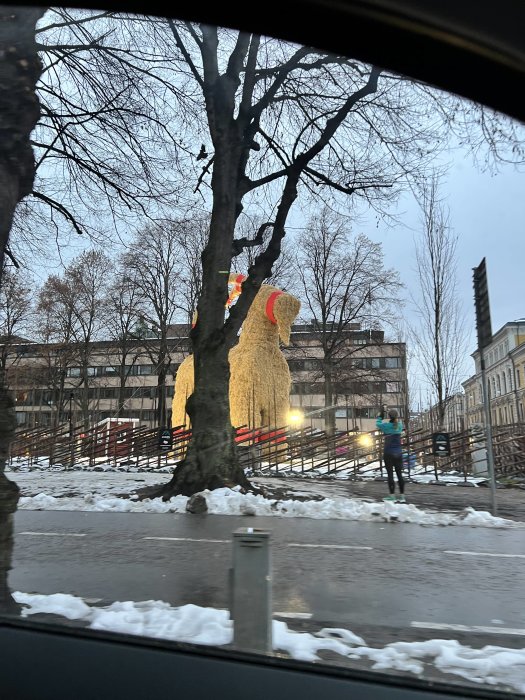 Stort halmgubbe-skulptur, vinter, snörester, stadsmiljö, person joggar, träd utan löv, dämpad belysning.