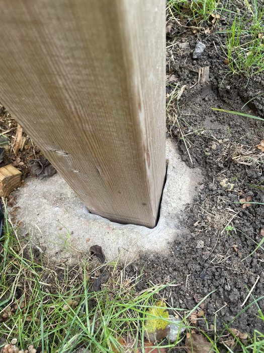 Trästaketstolpe vid betongkant omgiven av gräs och jord.