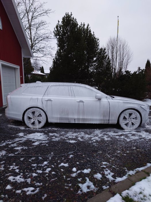 Bil täckt av snö och is, parkerad utanför rött hus, svensk flagga i bakgrunden, gråmulet väder.