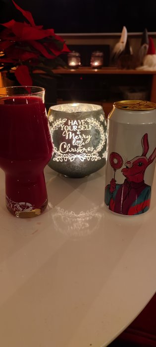Röd dryck i glas, burk med kaninmotiv, juldekoration, ljus med text "Merry little Christmas", mysig atmosfär.