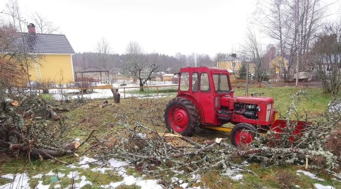 Röd traktor, nedfallna grenar, snöfläckar, gul stuga, landsbygdsmiljö.